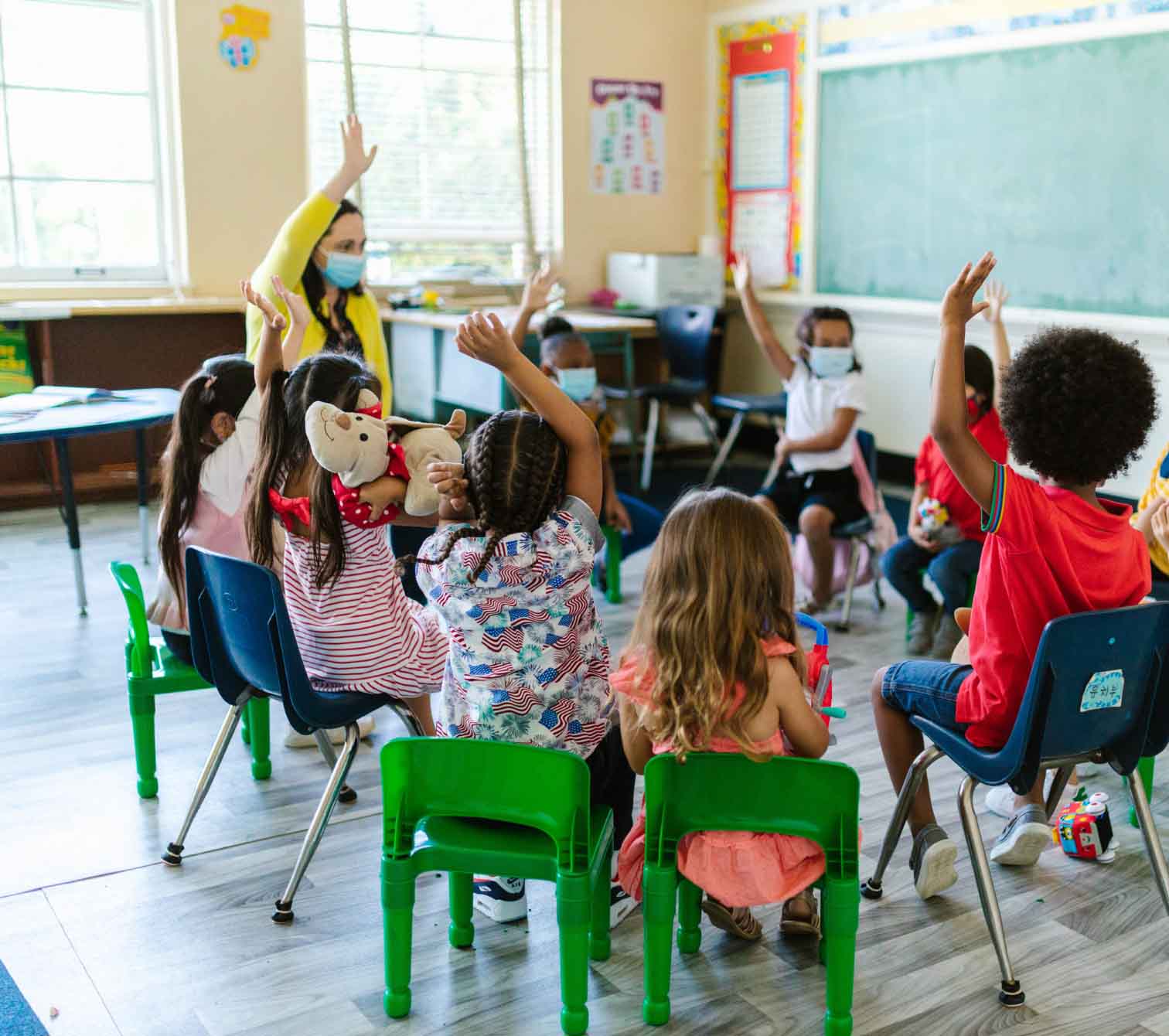 Elementary school students sit in a circle and some of them have their hands raised.